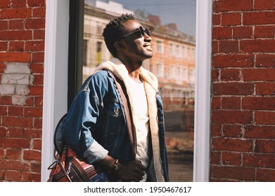 Portrait Smiling Young African Man Wearing A Jeans Jacket And Backpack Looking Up At Sunlight While Walking On A City Street Over Brick Wall Background