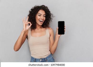 Portrait of a smiling young african woman holding blank screen mobile phone and showing ok over gray background