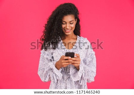 Portrait of a smiling young african woman in summer dress standing isolated over pink background, using mobile phone
