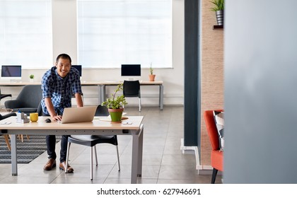 Portrait Of A Smiling Young Asian Designer Working On A Laptop While Leaning On His Desk In A Large Modern Office