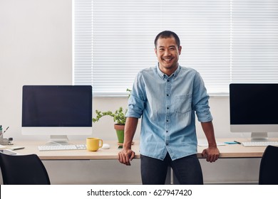 Portrait Of A Smiling Young Asian Designer Leaning On A Computer Table While Standing Alone In A Modern Office