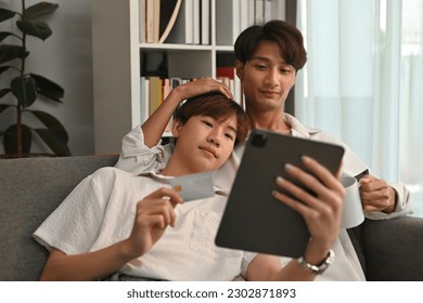 Portrait with Smiling of Young Asian gay couple using tablet and holding credit card while embracing and sitting on sofa at home. - Powered by Shutterstock