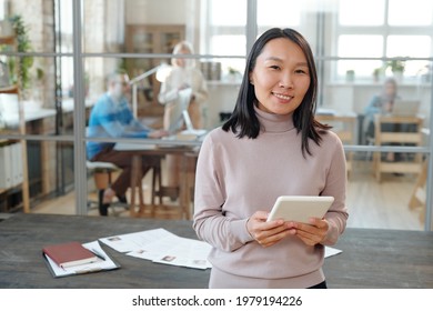 Portrait of smiling young Asian HR manager with tablet standing against wooden table with resumes of candidates