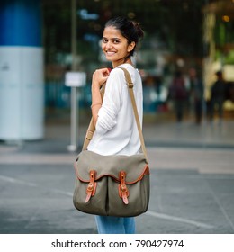 Portrait Of A Smiling Young Asian Indian Woman Standing And Looking Over Her Shoulder In The Day In The City. She Has A Green Sling Bag Over Her Shoulder.