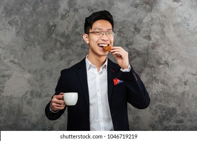 Portrait of a smiling young asian man dressed in suit drinking coffee and eating cookie over gray background