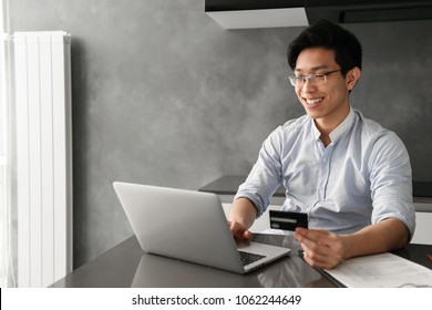 Portrait Of A Smiling Young Asian Man Holding Plastic Credit Card While Sitting At The Table With Laptop Computer At Home