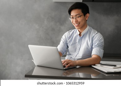 Portrait Of A Smiling Young Asian Man Working On Laptop Computer While Sitting At The Table With Paperwork At Home