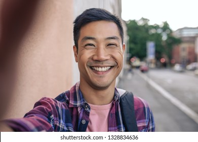 Portrait Of A Smiling Young Asian Man Standing On A Sidewalk In The City Taking A Selfie