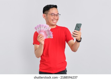 Portrait Of Smiling Young Asian Man Using Mobile Phone And Holding Money Banknotes Isolated Over White Background