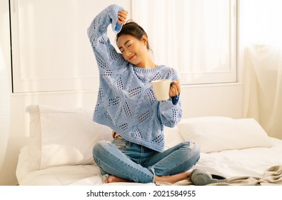 Portrait Of Smiling Young Asian Woman Wearing Knitted Warm Sweater And Holding Cup Of Coffee Or Tea While Relaxing Sitting On Bed In The Morning At Home, Lazy Day Off Concept.