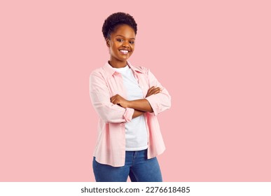 Portrait of smiling young attractive african american woman with hands folded on the chest in pink shirt, jeans and white t-shirt standing half turned isolated on pink background looking at camera. - Powered by Shutterstock