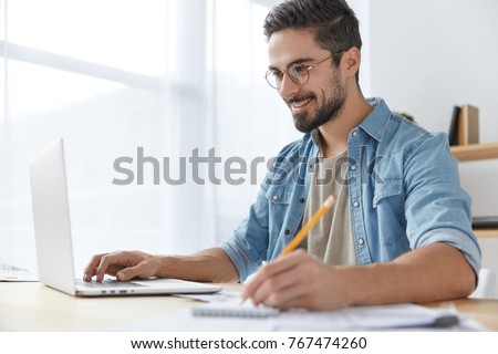 Portrait of smiling young attractive male student with trendy hairdo, wears glasses, makes notes, writes information from laptop computer, prepares for presenting his scientific work, works at home