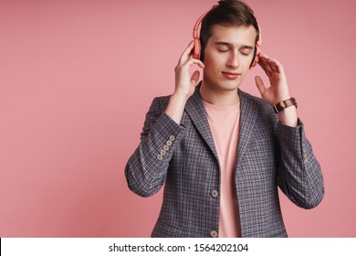 Portrait of a smiling young attractive man wearing jacket standing isolated over pink background, lstening to music with wireless headphones