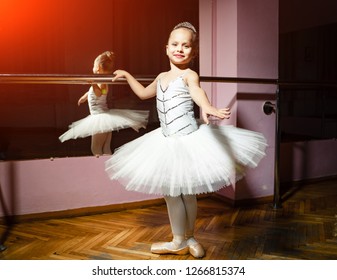 Portrait Of Smiling Young Ballerina In White Tutu And Pointes Posing Isolated In Dance Studio. Small Balet Dancer Standing Near Bar And Mirror, Preparing For Perfomance.