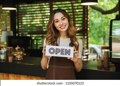 Portrait of a smiling young barista girl in apron holding open sign board while standing at the cafe - Powered by Shutterstock