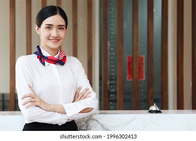 Portrait Of Smiling Young Beautiful Asian Female Receptionist Confidential Standing With Arms Crossed At Hotel Reception Counter Desk, Check In Hotel Service On Vacation Concept.  