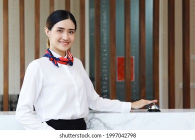 Portrait Of Smiling Young Beautiful Asian Female Receptionist Reaching Hand To Ring Silver Bell On Hotel Reception Service Desk, Check In Hotel Service On Vacation Concept.
