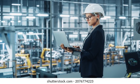 Portrait Of A Smiling Young Beautiful Female Engineer Wearing White Hard Hat, Using Laptop Computer In Office At Car Assembly Plant. Industrial Specialist Working On Vehicle Design In Modern Facility.