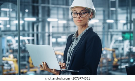 Portrait Of A Smiling Young Beautiful Female Engineer Wearing White Hard Hat, Using Laptop Computer In Office At Car Assembly Plant. Industrial Specialist Working On Vehicle Design In Modern Facility.