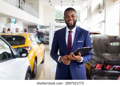 Portrait Of Smiling Young Black Car Dealer In Suit At Workplace In Showroom, Cheerful African American Salesman In Auto Dealership Office Standing Near New Vehicles, Waiting For Customers, Copy Space