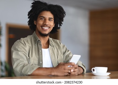 Portrait Of Smiling Young Black Guy Relaxing With Smartphone At Table In Cafe, Happy Millennial African American Man Drinking Coffee And Using Mobile Phone For Online Communication, Copy Space - Powered by Shutterstock