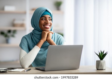 Portrait Of Smiling Young Black Islamic Woman In Hijab Sitting At Desk With Computer And Looking Away, Dreamy African Muslim Lady In Headscarf Enjoying Freelance Work In Home Office, Copy Space
