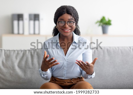 Portrait of smiling young black psychologist gesturing and smiling at camera, sitting on comfy couch at modern office. Positive African American marriage counselor on psychotherapy session