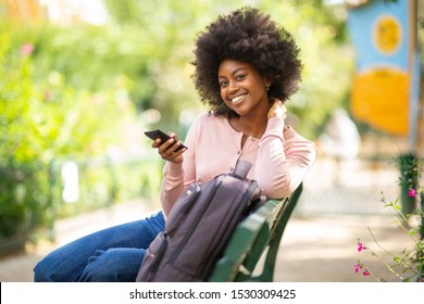 Portrait Of Smiling Young Black Woman Sitting On Park Bench Holding Mobile Phone