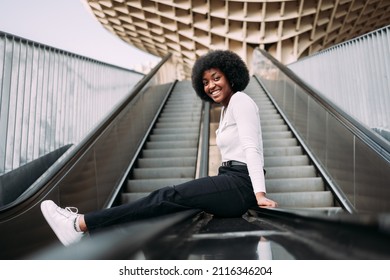 Portrait Of Smiling Young Black Woman On Top Of Escalator Railing Looking At Camera In Profile