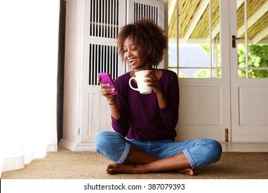 Portrait Of A Smiling Young Black Woman Sitting On Floor At Home With Cell Phone