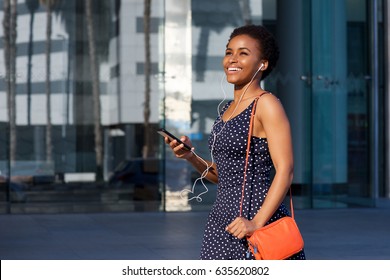 Portrait Of Smiling Young Black Woman Walking With Earphones And Mobile Phone