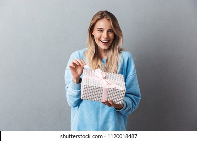 Portrait Of A Smiling Young Blonde Girl Unwrapping Present Box Isolated Over Gray Background