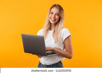 Portrait of a smiling young blonde girl holding laptop computer isolated over yellow background