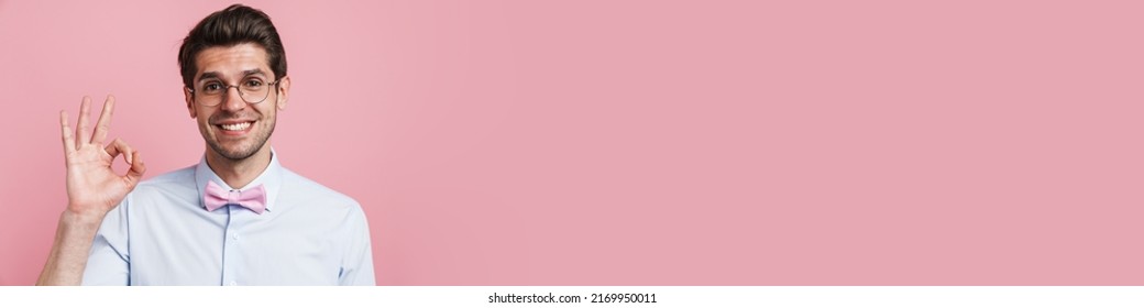 Portrait Of A Smiling Young Brunette Nerd Man Wearing Shirt And Bowtie Standing Over Pink Wall Background Showing Ok