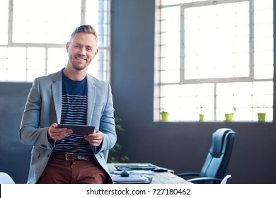 Portrait Of A Smiling Young Businessman Leaning On His Desk In A Large Modern Office Working Online With A Digital Tablet