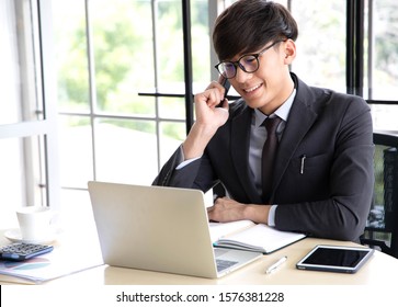 Portrait of a smiling young businessman  talking on his smartphone while he working on his computer, sitting in his office at desk wearing a black suit. - Shutterstock ID 1576381228