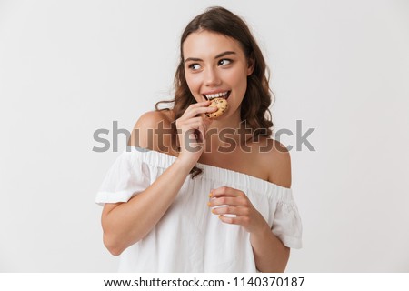 Portrait of a smiling young casual brunette woman eating a cookie isolated over white background
