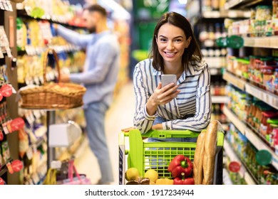 Portrait Of Smiling Young Casual Woman Leaning On Shopping Trolley Cart Indoors, Using Her Mobile Phone, Posing And Looking At Camera. Satisfied Female Customer Buying Products In Local Grocery Store