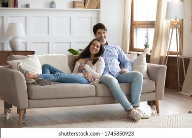 Portrait of smiling young caucasian couple sit relax on comfortable sofa in design renovated living room, happy renters tenants cuddle rest on couch at home, hugging and embracing in own apartment