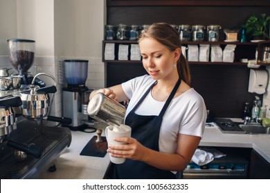 Portrait of smiling young Caucasian woman barista  in apron pouring milk in coffee. Business owner concept. 
