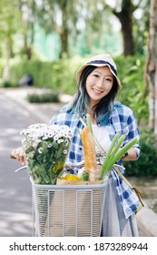 Portrait Of Smiling Young Chinese Woman Riding On Bicycle With Bouquet Of Flowers And Groceries Bag In Basket