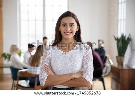 Portrait of smiling young company employee, standing in modern office with colleagues on background. Happy attractive millennial corporate worker manager looking at camera, posing for photo.