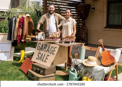 Portrait Of Smiling Young Couple Standing At Table With Garage Sale Sign And Selling Stuff In Backyard