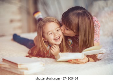 Portrait of a smiling young cute mother and daughter reading a book lying and relax in the bed in a bright big white room 