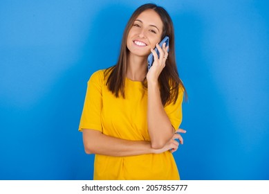 Portrait Of A Smiling Young European Brunette Woman Wearing Yellow T-shirt On Blue Background Talking On Mobile Phone. Business, Confidence And Communication Concept.