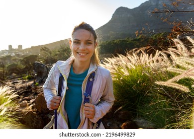 Portrait Of Smiling Young Female Hiker With Backpack Standing By Plants Against Mountain And Clear Sky During Sunrise