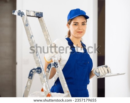 Portrait of smiling young female plasterer wearing blue overalls and cap standing with finishing trowel in hands near step ladder inside building under construction