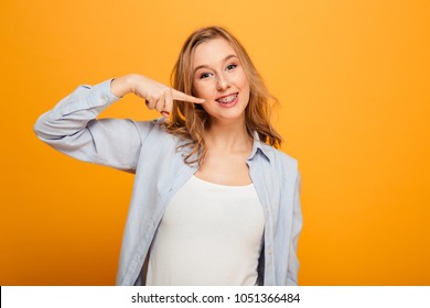 Portrait of a smiling young girl with braces pointing finger at her mouth isolated over yellow background