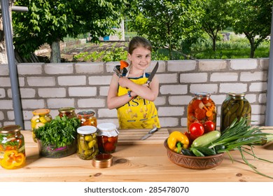 Portrait Of Smiling Young Girl Holding Canning Supplies At Wooden Table Covered With Fresh Vegetables And Jars Of Pickled Preserves Outdoors By Brick Garden Wall