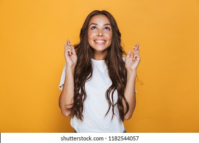 Portrait of a smiling young girl with long brunette hair standing over yellow background, holding fingers crossed for good luck